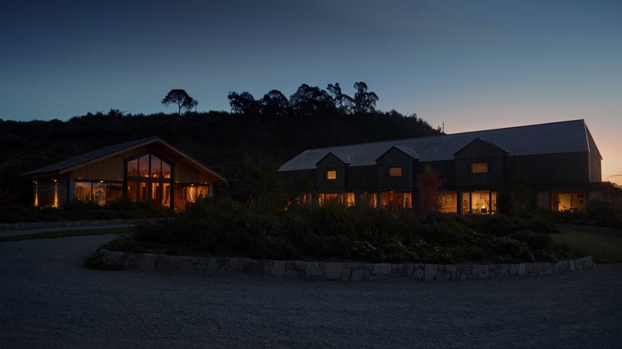 A main residence and a detached guest structure at dusk, both lit from within across a gravel motor court