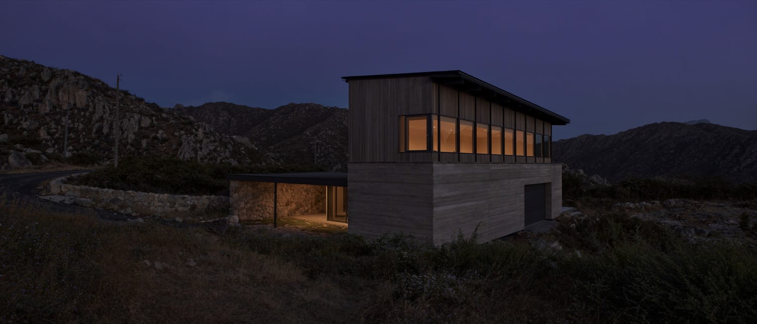 A contemporary custom home at blue hour, warm interior light behind deep glazing, set into a foothill landscape
