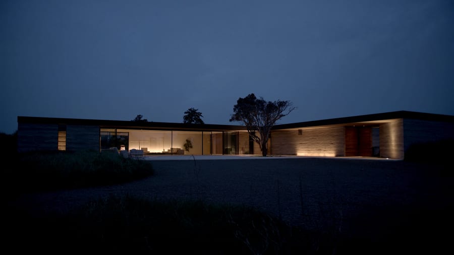 A long low custom home in the evening, warm light returning through deep glazing onto a gravel court