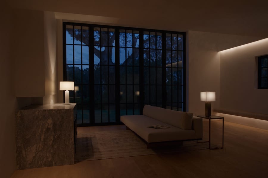 A quiet living room at dusk lit only by two table lamps and a cove light, tall steel-framed glazing holding the last blue of the sky