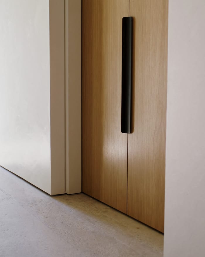 A flush timber utility-room door with a blackened-steel pull, meeting a limestone floor at a clean shadow gap